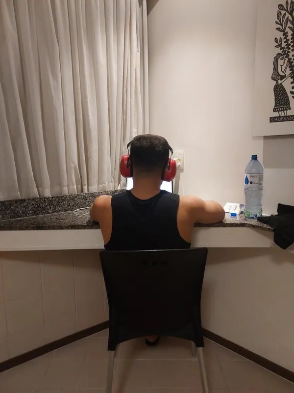 Avimar de costas, com abafador vermelho no ouvido, estudando numa mesa improvisada de quarto de hotel.