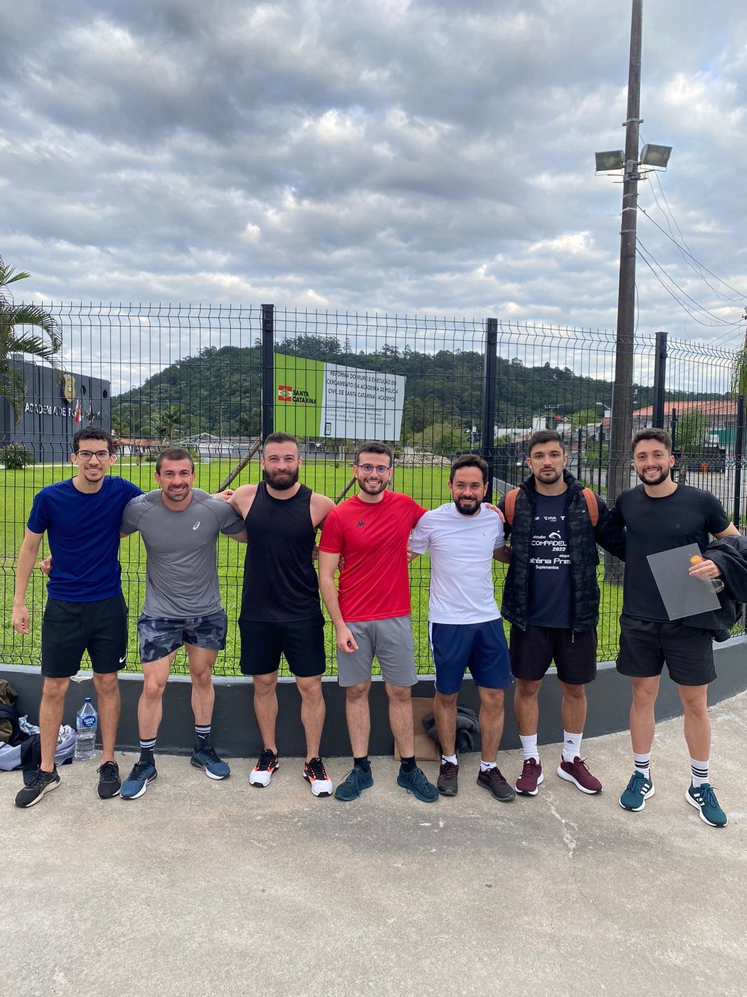 Grupo de sete candidatos após o teste físico, diante da placa da Academia de Polícia Civil de Santa Catarina.