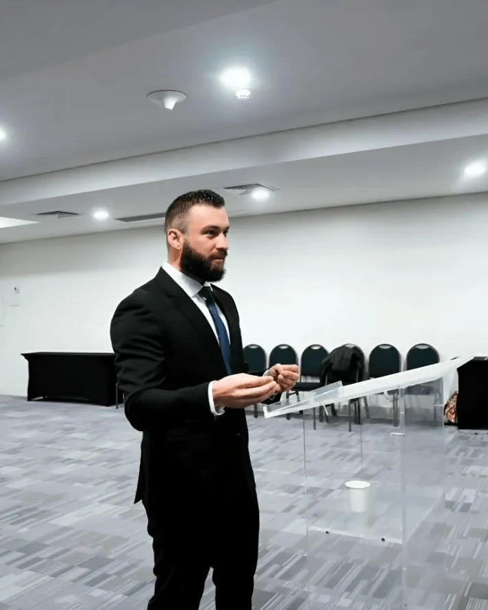 Avimar de terno preto e gravata, em pé com gesto de fala diante de um púlpito de vidro, na sala de um simulado de prova oral com cadeiras e mesa ao fundo.